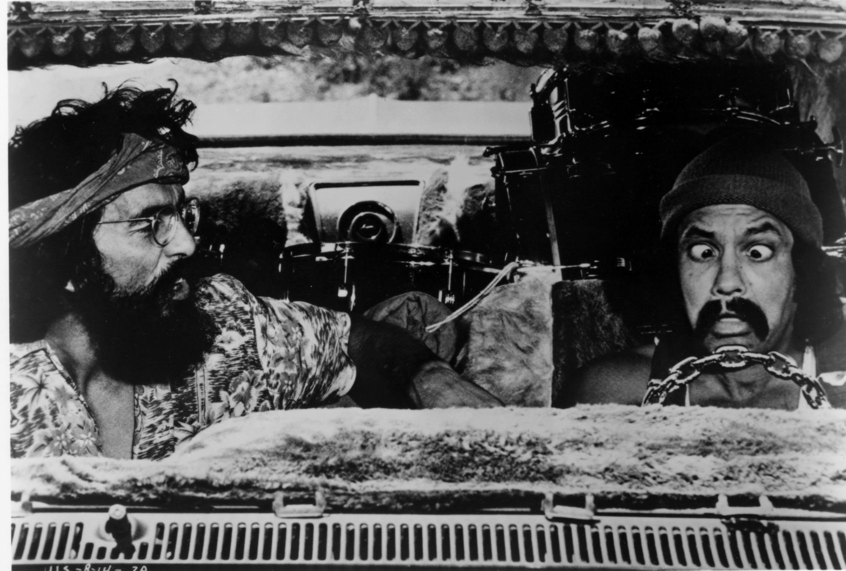 Black and white photo still of two men seen through a van's windshield. One is bearded with a bandana, who's looking at the other man, who is sitting cross-eyed in front of a steering wheel made of a chain