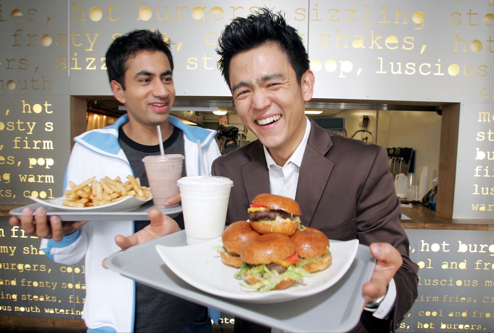 Two men are smiling and laughing. One holds a tray with french fries and a chocolate shake while the other holds a tray with 4 hamburger slider and a white takeout cup